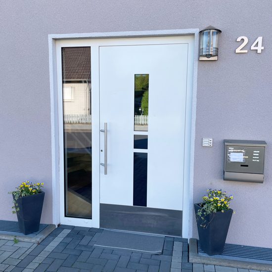 Eingang WG Helle Eingangstür mit Glaselementen und Hausnummer 24, umgeben von Blumentöpfen.