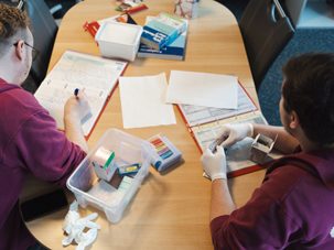 Zwei Personen arbeiten an einem Tisch mit medizinischen Materialien und Dokumenten.