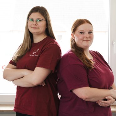 Aktuelle Auszubildende Zwei Frauen mit langen Haaren, stehen nebeneinander, beide in bordeauxroten T-Shirts.