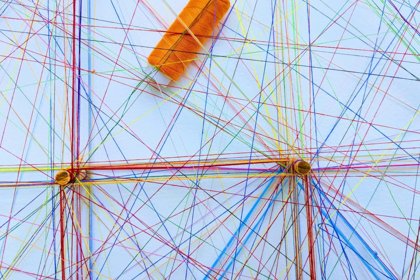 Bunte Fäden kreuzen sich auf einer weißen Fläche, mit einer orangefarbenen Garnrolle.