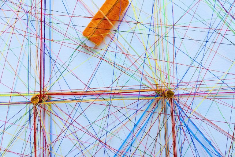 Kooperation Bunte Fäden kreuzen sich auf einer weißen Fläche, mit einer orangefarbenen Garnrolle.