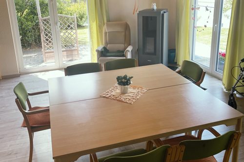 Wohngemeinschaften Holztisch mit Stühlen, Pflanze in der Mitte, helles Zimmer mit großen Fenstern und Gartenblick.